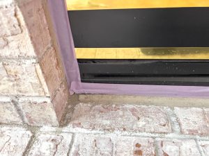 Close view of a brick fireplace with purple painter tape framing the black metal opening. The scene indicates paint preparation during renovation.